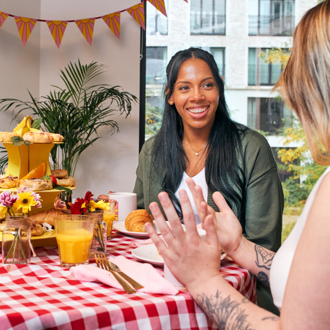 A table set with a red-and-white checkered cloth, featuring a tiered yellow stand filled with pastries and decorated with flowers. Glasses of orange juice, mugs, croissants, and slices of bread are arranged around the table. Two people are seated, engaged in conversation, with a plant and window in the background and colourful bunting hanging above.