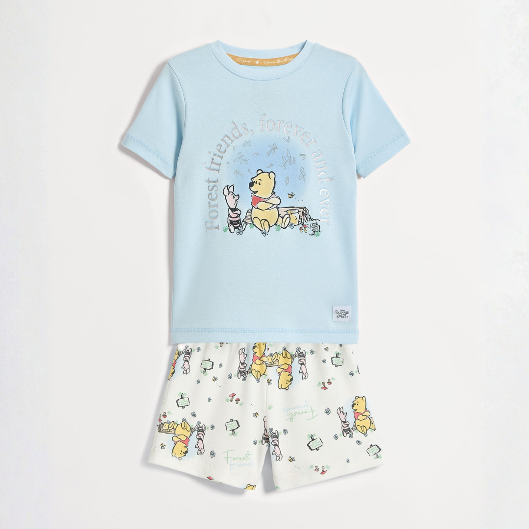 A light blue short‑sleeved pyjama T‑shirt featuring an illustration of Winnie the Pooh and Piglet sitting together, with the words “Forest friends, forever and ever” above them. The shirt is paired with white shorts patterned with small repeating illustrations of Winnie the Pooh, Piglet, honey pots, and flowers.