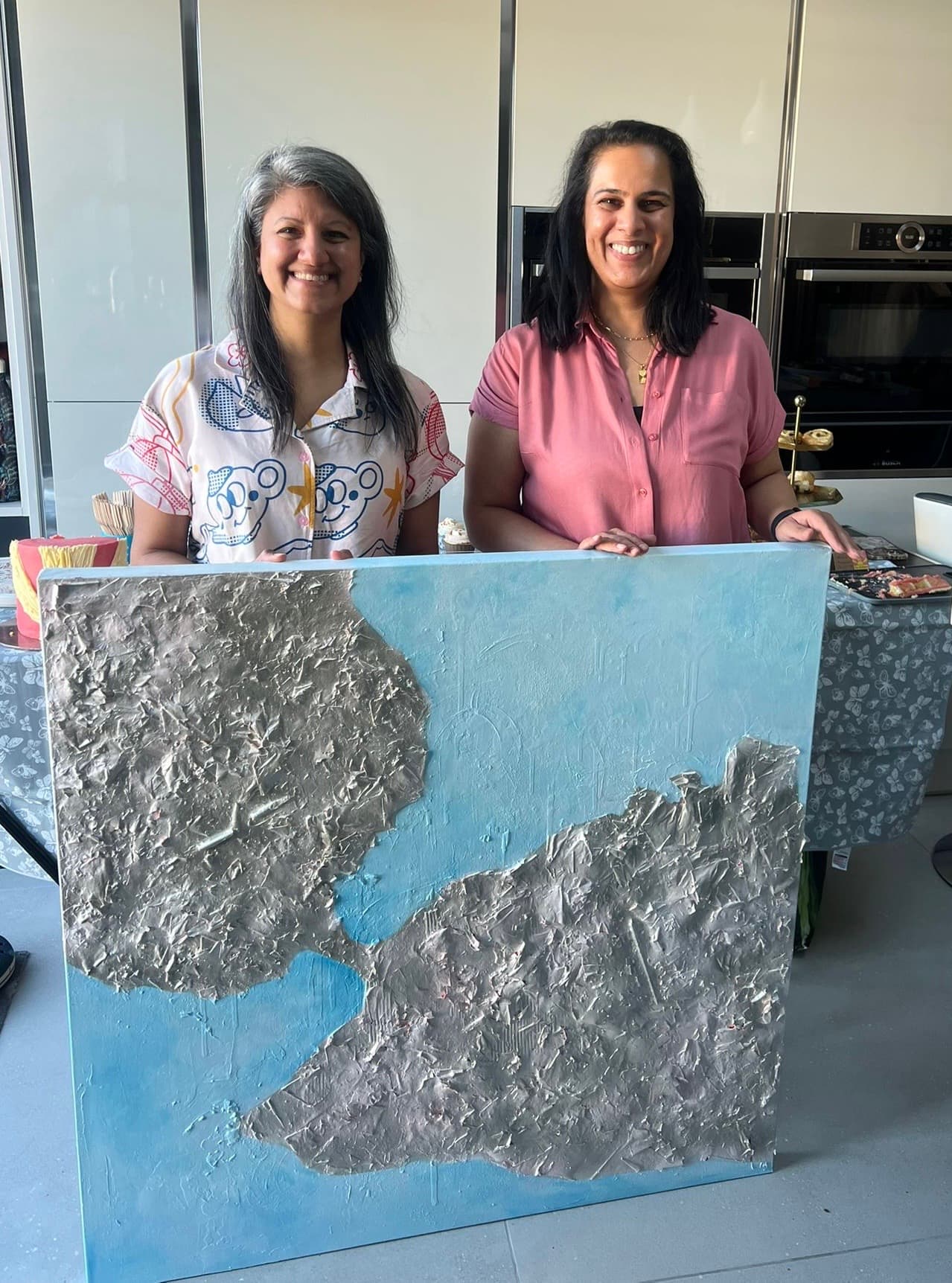 Two people stand behind a large abstract artwork featuring textured silver shapes on a light blue background in a modern indoor setting. The artwork is displayed in the foreground, while a table with cakes and refreshments can be seen behind it. The space includes contemporary kitchen units and appliances.
