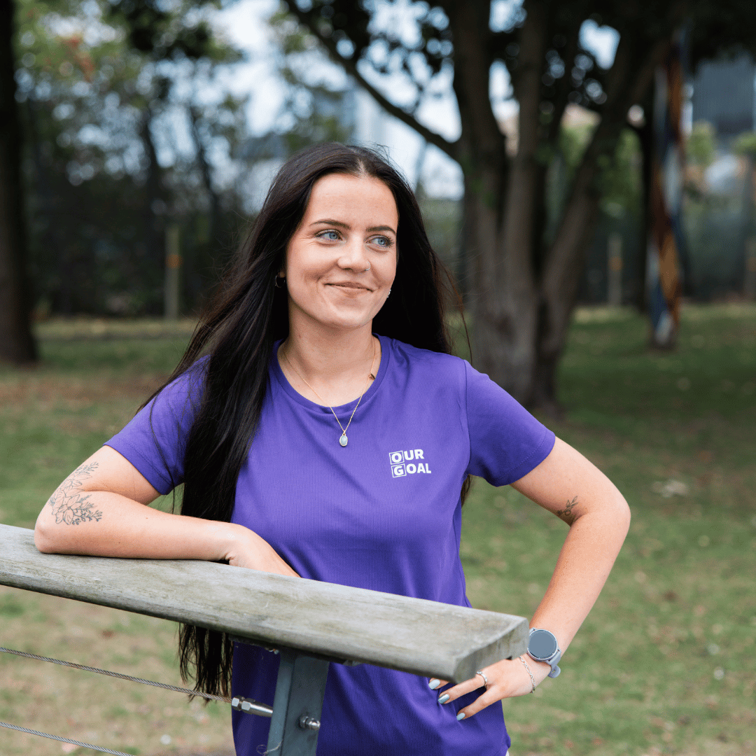 Image of a young women with long dark hair, looking into the distance wearing a purple T-Shirt.
