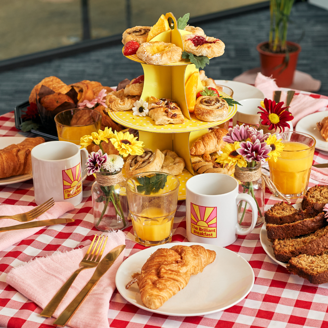 Close-up of a breakfast table with a red-and-white checkered cloth. A yellow tiered stand holds assorted pastries and is surrounded by plates with croissants, mugs, glasses of orange juice, and small flower arrangements.