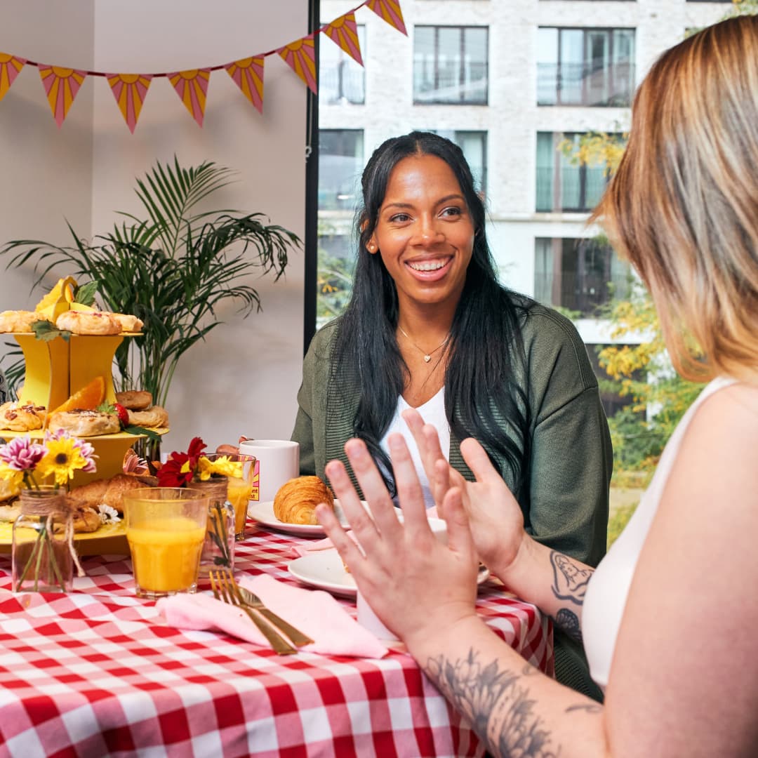 Two people chatting over breakfast at a table filled with pastries, fruit and drinks, decorated for a fundraising event.