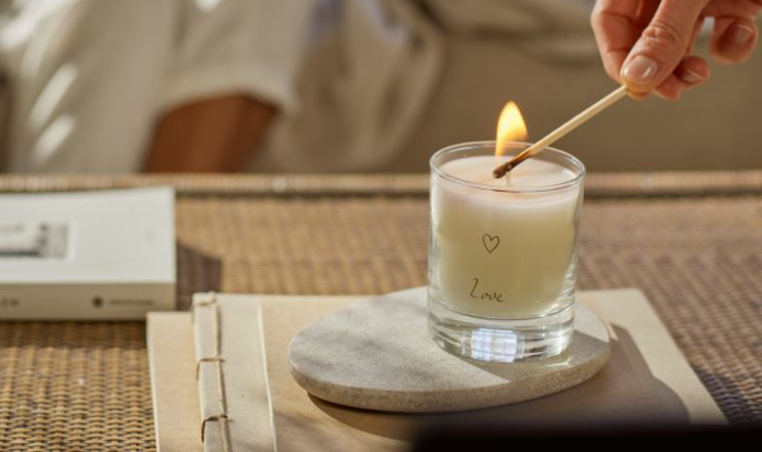 A hand lighting a white candle labeled ‘Love’ with a matchstick, placed on a round stone coaster atop a notebook on a woven surface, with a book in the background.