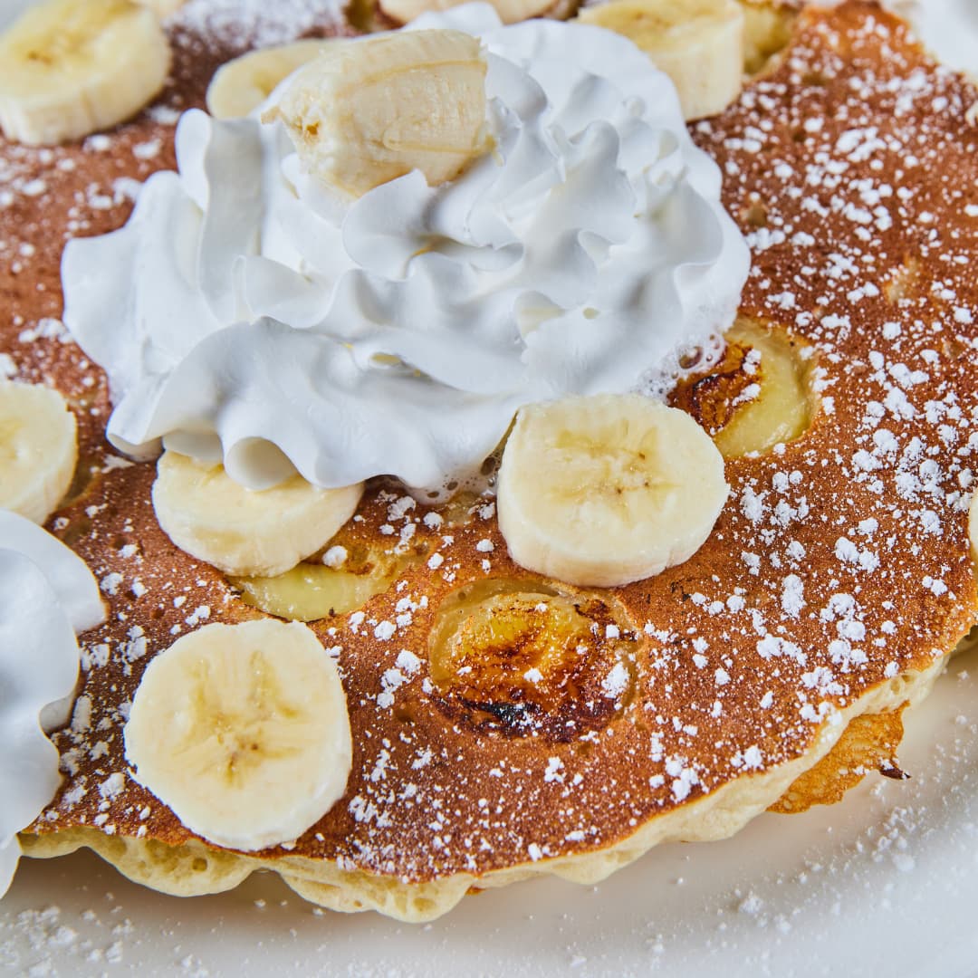 Close-up of golden-brown pancakes topped with sliced bananas, a generous swirl of whipped cream and a dusting of icing sugar. The pancakes are garnished with caramelised banana slices and served on a white plate.