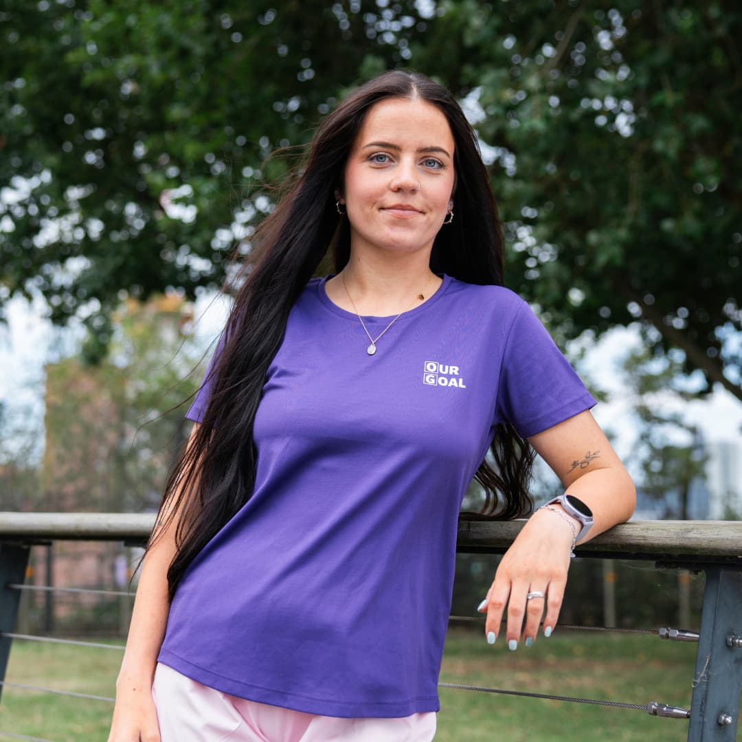 A young woman with long dark hair wearing a purple 'Our Goal' branded T-shirt, leaning on a railing in an outdoor park setting with trees in the background.