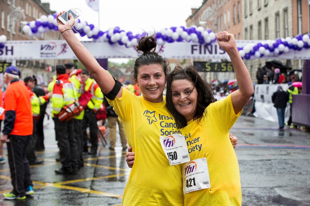 Two women in yellow Irish Cancer Society t-shits smile and raise their arms in celebration at a VHI race finish line with men and passers by in their background.