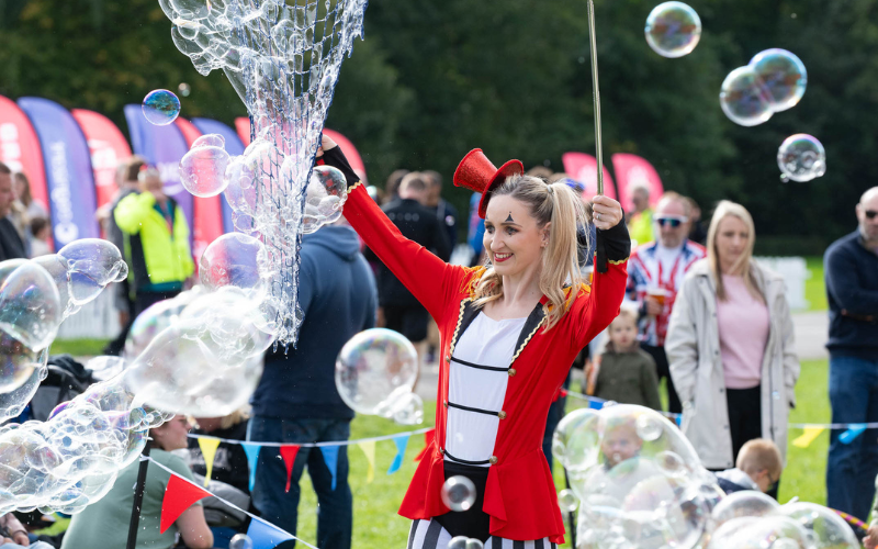 A woman is wearing fancy dress with a red top hat and a red tunic. She is smiling and holding a wand wand is making large bubbles. There are people and families in the background.