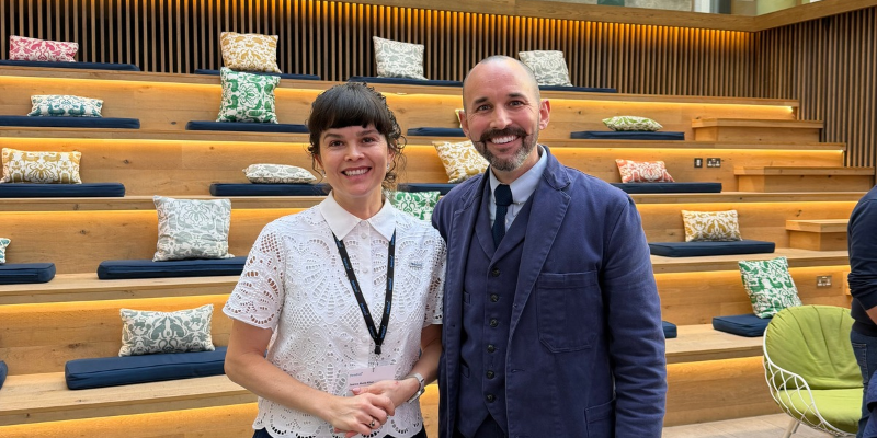 Two people stand side by side in a modern indoor venue with tiered wooden seating behind them. The seating area features navy cushions and patterned decorative pillows, with warm lighting built into each tier. Plants in rectangular planters line the upper level. One person wears a white short-sleeved patterned blouse and a dark skirt, while the other wears a blue suit and tie. The space has a light stone floor and contemporary furnishings.