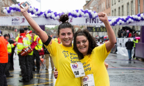 Two women in yellow Irish Cancer Society t-shits smile and raise their arms in celebration at a VHI race finish line with men and passers by in their background.