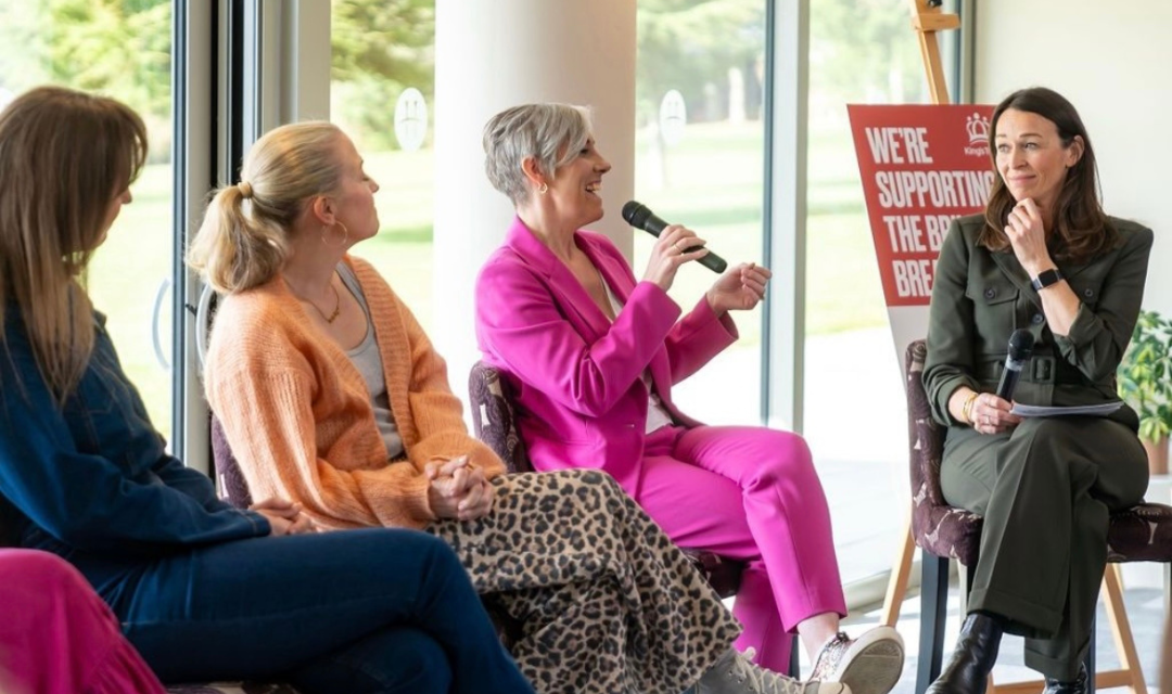Panel discussion taking place indoors with four people seated on chairs. One person in a bright pink suit is speaking into a microphone, while another holds notes and a microphone. A red sign in the background reads ‘We’re Supporting The Brilliant Breakfast.’ Large windows show greenery outside.
