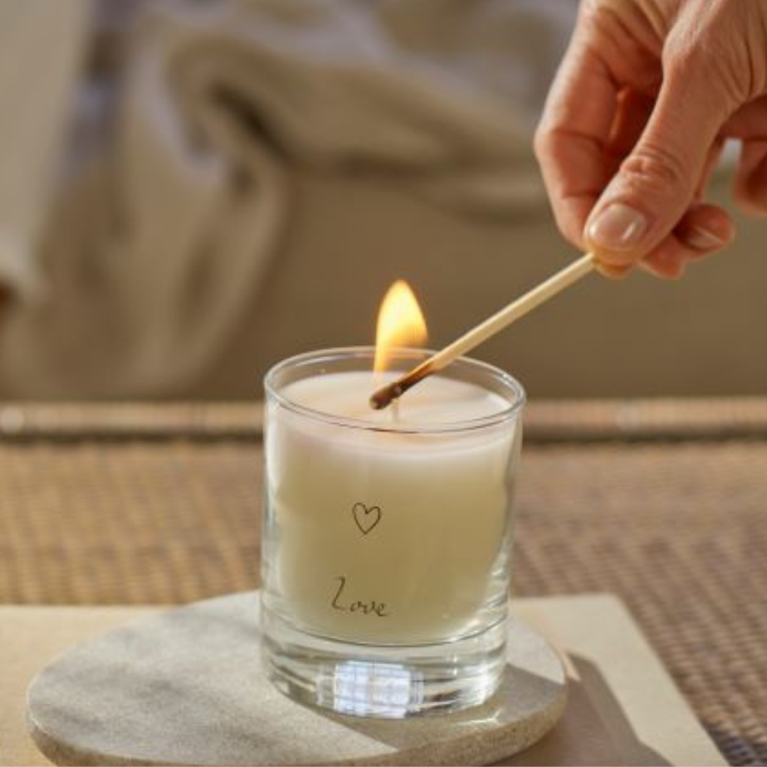 A hand lighting a white candle labeled ‘Love’ with a matchstick, placed on a round stone coaster atop a notebook on a woven surface, with a book in the background.