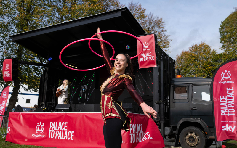 A women in fancy dress is spinning two hula hoops with her hand raised in the air. Behind her, a female is playing guitar and singing on a stage with 'King's Trust Palace to Palace' branding.
