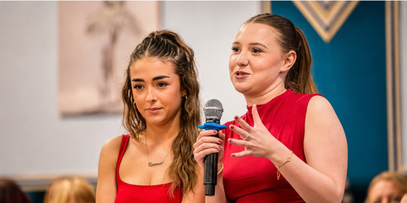 Two young women stand at a lectern during a Brilliant Breakfast fundraising event, with one speaking into a microphone while attendees listen in the background.