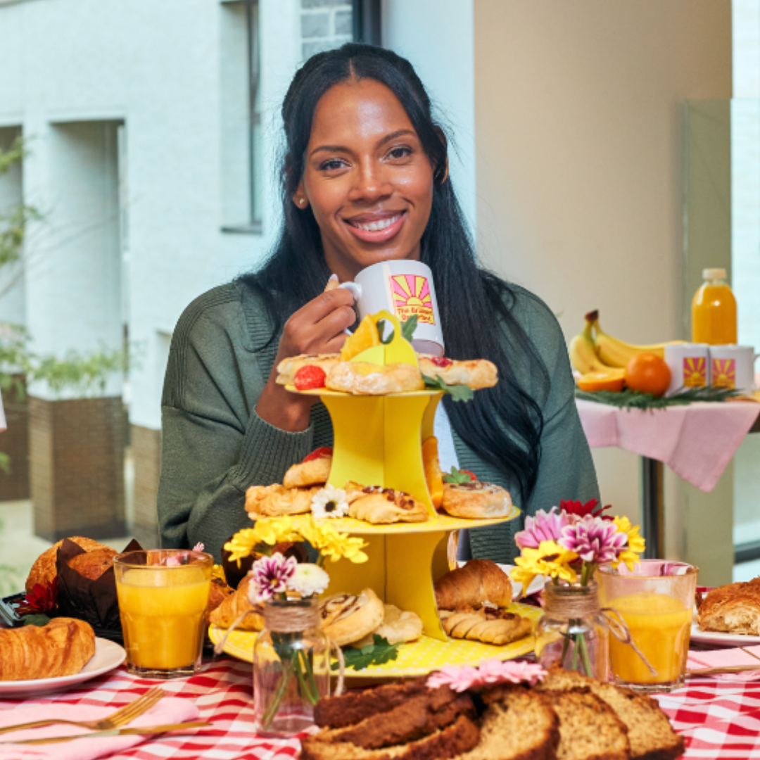 A young woman sat at a table with a red-and-white checkered cloth, holding a mug with The Brilliant Breakfast logo. The table is decorated with a yellow tiered stand of pastries, fresh flowers, croissants, and glasses of orange juice. A window view and additional breakfast items are visible in the background.
