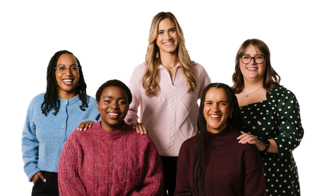 Five people standing together in two rows against a white background, wearing casual clothing in various colours including blue, pink, green with white polka dots, maroon, and burgundy. Two individuals are seated in front while three stand behind them, with hands resting gently on shoulders.