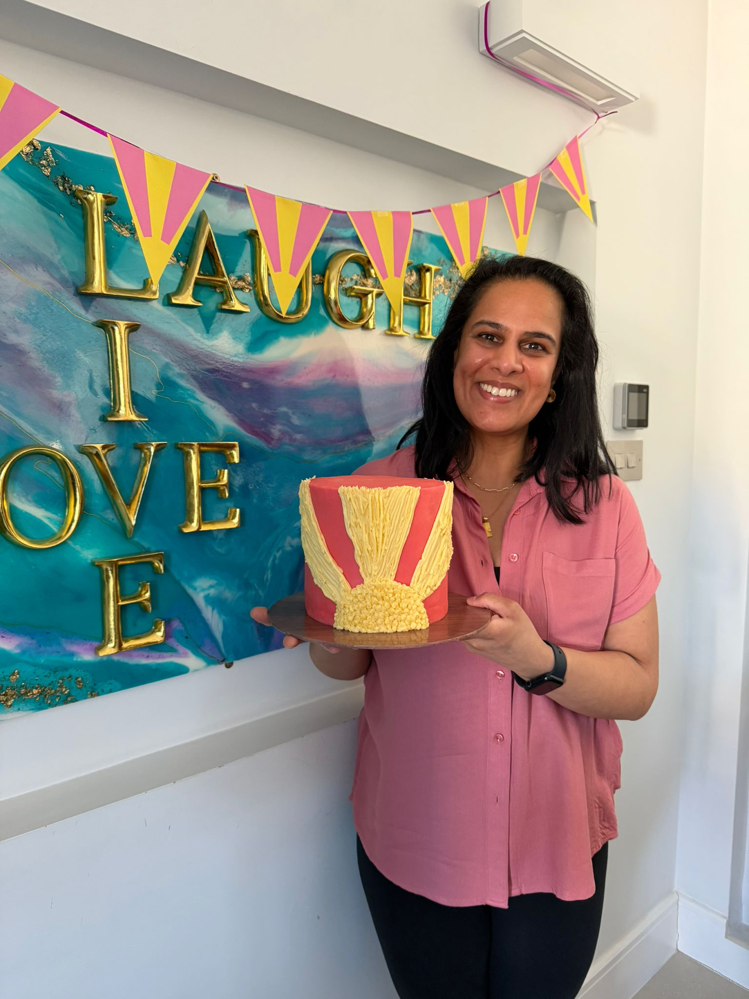 A person stands indoors holding a pink cake decorated with yellow icing in the shape of a sun rays. Behind them is a colourful wall artwork in shades of blue, purple and turquoise with large gold letters. Pink and yellow bunting hangs above the artwork, creating a celebratory atmosphere.