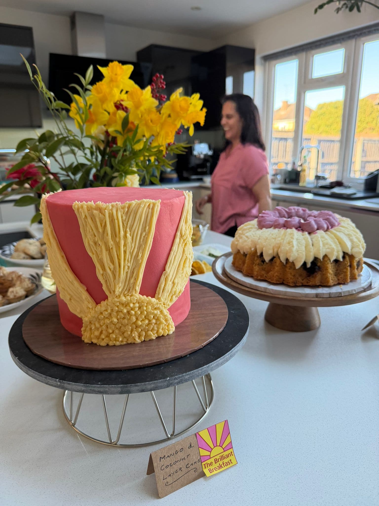 A pink cake decorated with yellow icing in the shape of sun rays sits on a cake stand in the foreground of a kitchen. Another iced cake is displayed on a wooden stand beside it, while a vase of bright yellow daffodils and a selection of baked goods are visible in the background. A small sign in front of the cake reads “The Brilliant Breakfast,” and a person stands near the kitchen counter behind the display.
