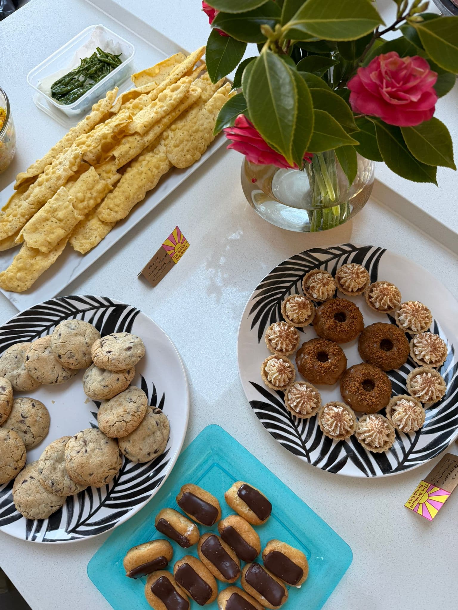 An assortment of homemade baked goods is displayed on a table at a fundraising event. Plates of chocolate chip cookies, iced mini cakes, ring-shaped cakes and chocolate-topped pastries surround a vase of pink flowers. A platter of savoury crackers and a small sign featuring The Brilliant Breakfast logo are also visible, creating an inviting breakfast spread.