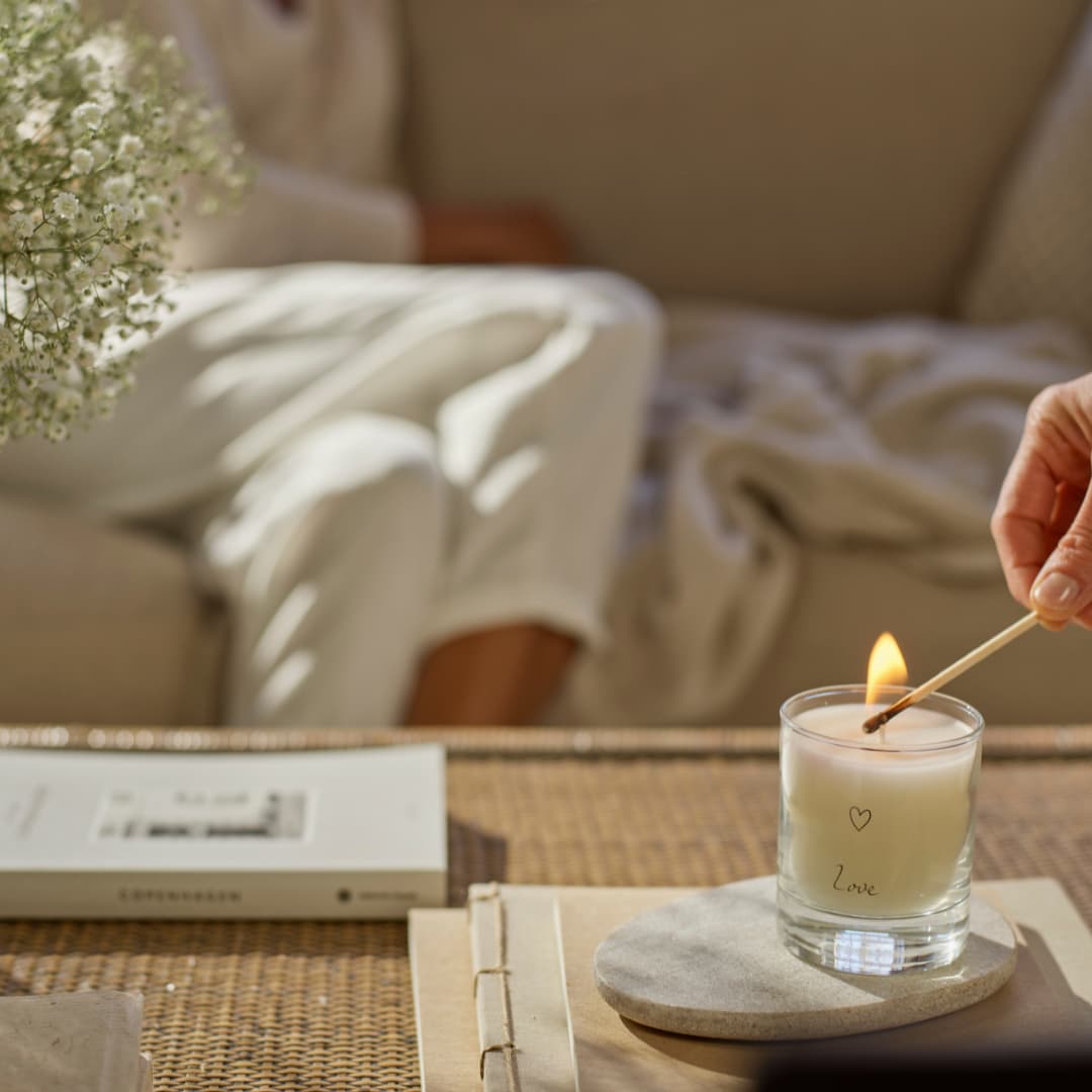Hand lighting a white candle in a glass holder on a marble coaster beside a book, creating a warm and relaxing home setting.