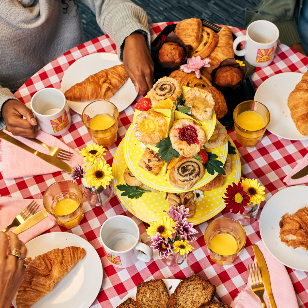 Close-up of a breakfast table with a red-and-white checkered cloth. A yellow tiered stand holds assorted pastries and is surrounded by plates with croissants, mugs, glasses of orange juice, and small flower arrangements.