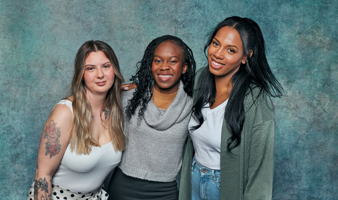 Three young women standing close together in front of a textured blue-green backdrop, with arms around each other. One person is wearing a white sleeveless top with a polka-dot skirt, another is in a grey cowl-neck sweater, and the third is in a white top with a green cardigan and jeans